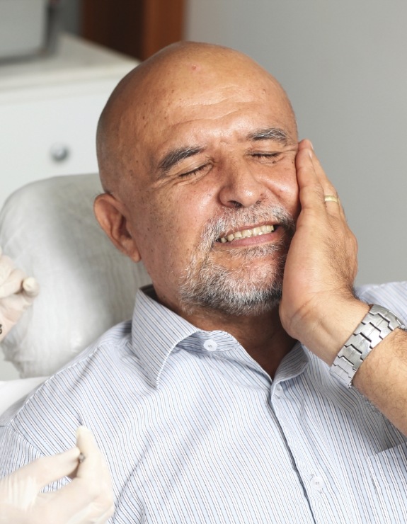 Older man in dental chair holding his cheek in pain