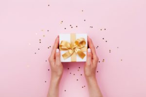 White gift box with gold ribbon being held by two hands with gold confetti sprinkled around them against a pink background 