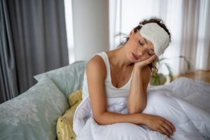 Woman wearing white shirt and sleep mask sitting in bed holding her hand over her jaw